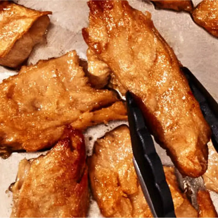 Golden-brown roasted Daring Plant-Based Unbreaded Gluten-Free Chicken Pieces on a baking sheet with black tongs holding one piece.