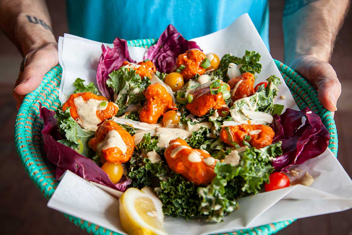 Two hands holding a teal colored straw basket filled with a plant based Caesar salad on parchment paper, topped with No Bones Frozen Cauliflower Wings in an orange colored sauce.