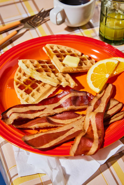 A red breakfast plate with a waffles, slices of oranges, and several pieces of Prime Roots Bacon