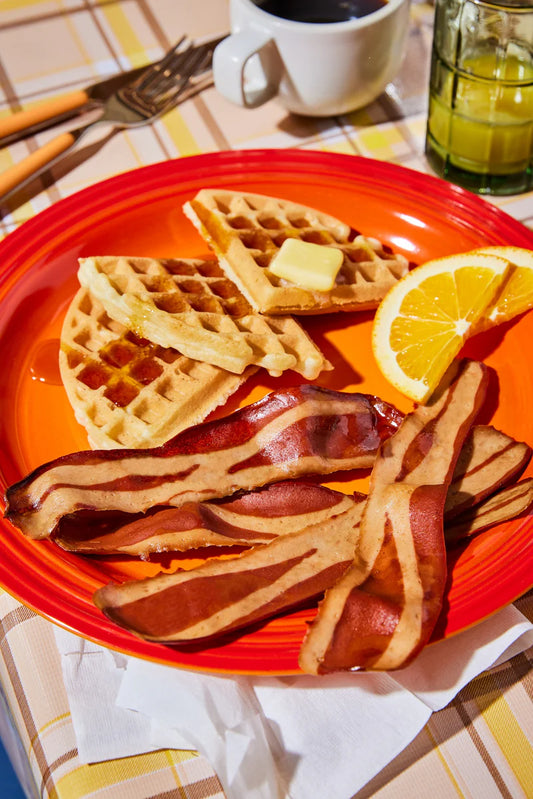 A red breakfast plate with a waffles, slices of oranges, and several pieces of Prime Roots Bacon
