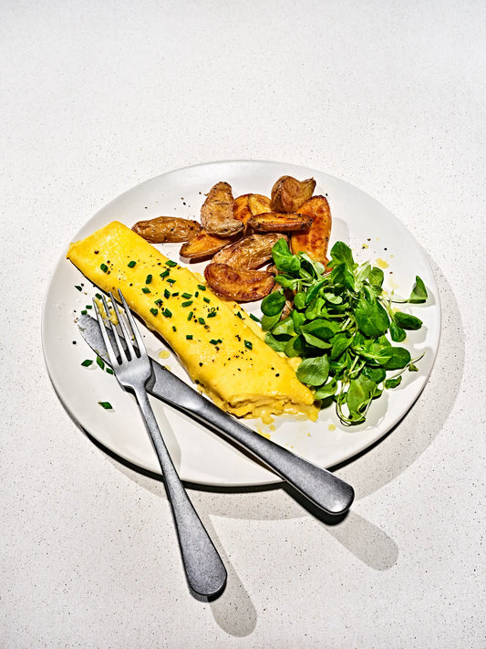A plated breakfast featuring a folded Just Egg omelette with herbs, roasted potato wedges, and green salad on a white plate with a fork and knife, set on a light speckled countertop.