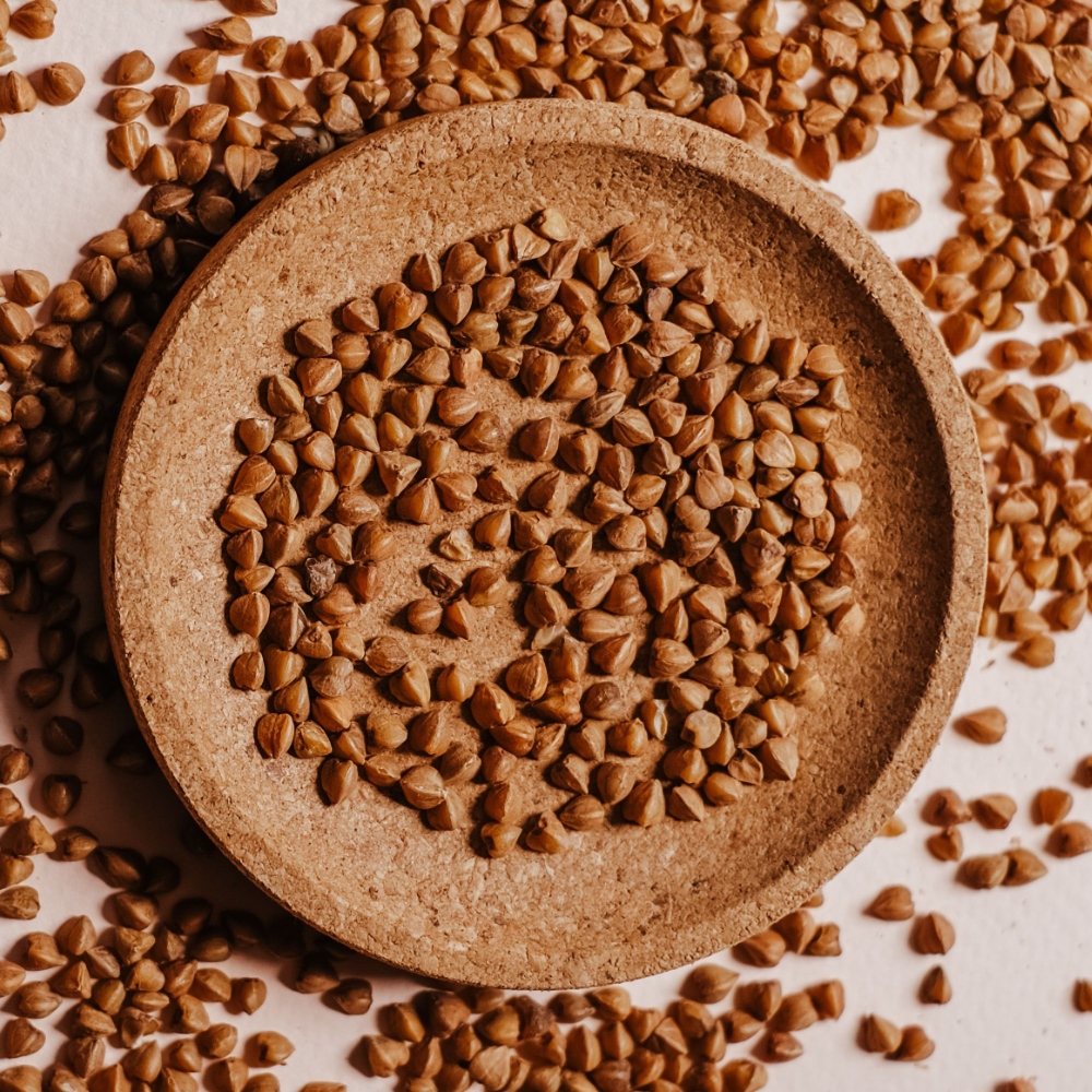 A top-down view of brown buckwheat groats arranged in a round beige ceramic dish, with additional scattered buckwheat seeds around the dish on a light background.