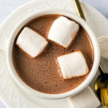 A top-down view of a white ceramic cup filled with hot chocolate, topped with three Dandies Classic Vegan Marshmallows. The cup sits on a matching white textured plate, with a gold spoon and a napkin partially visible on a light background.