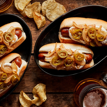 Two black oval platters holding Field Roast Plant Based Stadium Dog hot dogs topped with sliced green jalapeños, caramelized onions, and white sauce, arranged on a rustic wooden table with potato chips and beer glasses around them.