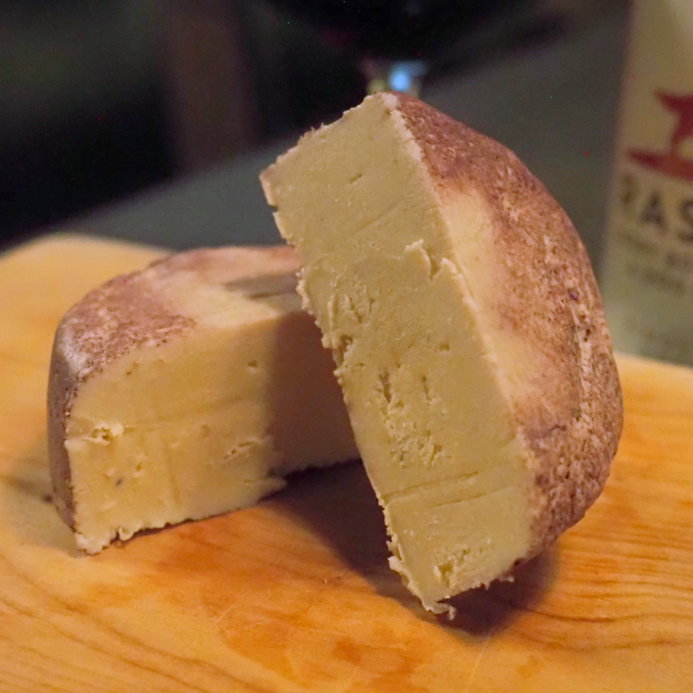 A close-up of a cut wheel of Rose City Creamery Perfect Pinot cheese on a wooden board, with one half leaning against the other. The cheese has a pale yellow interior and a red wine colored rind, set against a dark, blurred background with a wine bottle partially visible.