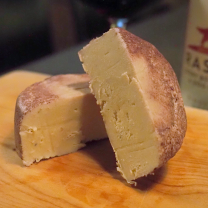 A close-up of a cut wheel of Rose City Creamery Perfect Pinot cheese on a wooden board, with one half leaning against the other. The cheese has a pale yellow interior and a red wine colored rind, set against a dark, blurred background with a wine bottle partially visible.