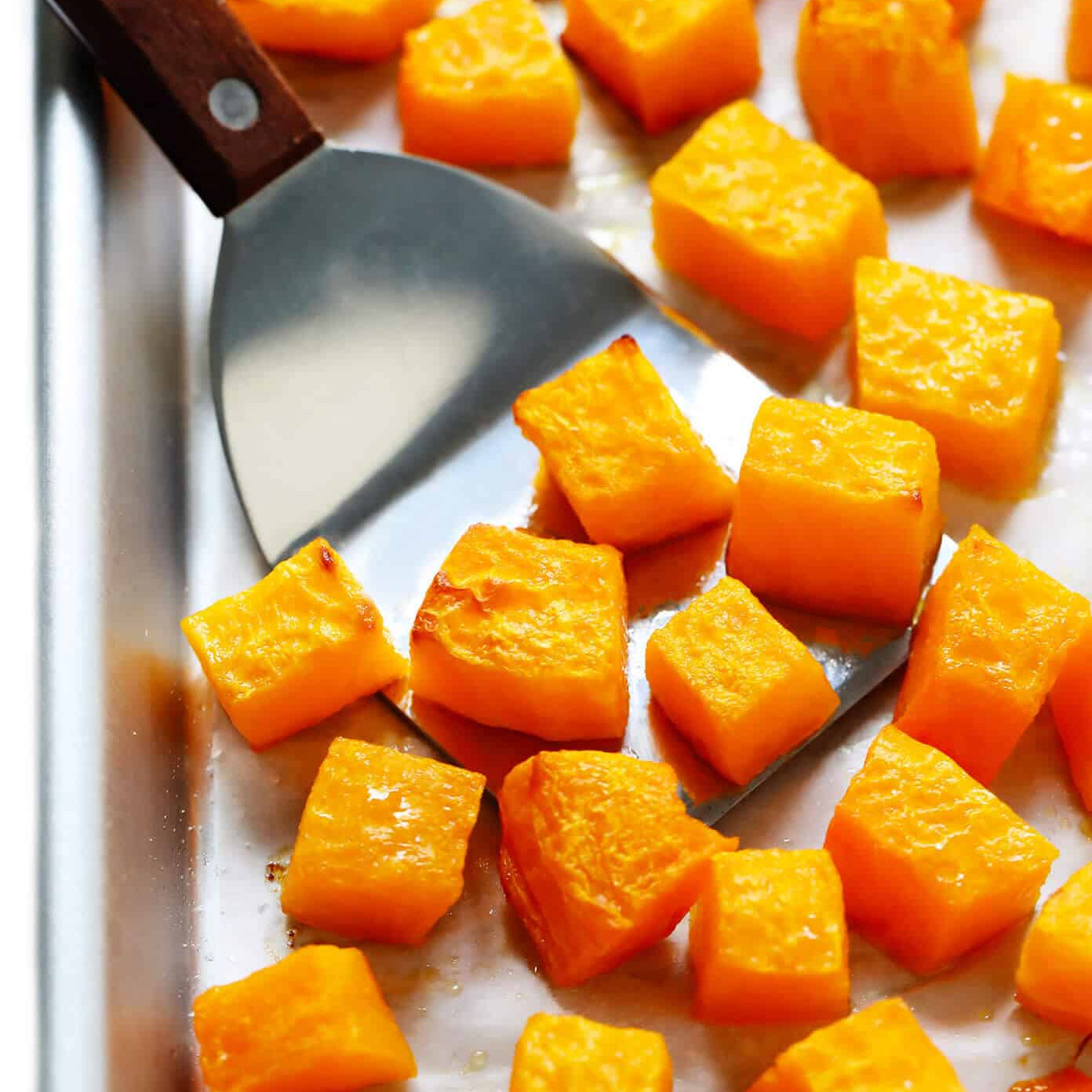 Roasted butternut squash cubes scattered on a baking sheet, with a metal spatula lifting one piece. The cubes are glossy and golden-orange, with some lightly browned edges.