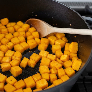 Cubed orange butternut squash pieces cooking in a black skillet with a wooden spoon on a stovetop.
