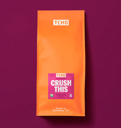 An orange stand-up pouch of TCHO Crush This cacao nibs on a solid dark magenta background. The package features a white TCHO logo at the top and a pink and white 'CRUSH THIS' label in the center.