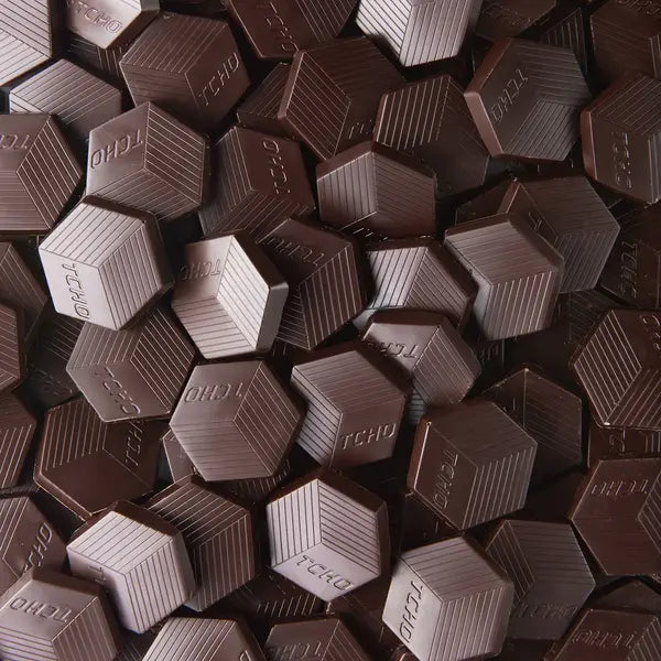 A close-up pile of hexagonal TCHO Hella Dark 81% chocolate pieces with the brand name 'TCHO' embossed on their faces.