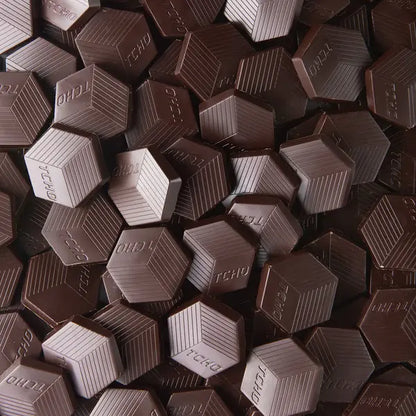 A close-up pile of hexagonal TCHO Hella Dark 81% chocolate pieces with the brand name 'TCHO' embossed on their faces.
