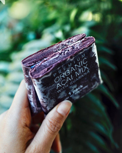 A hand holding an opened smoothie packet with frozen acai inside, labeled 'PREMIUM ORGANIC ACAI MIX WITH GUARANA' against a blurred green outdoor background.