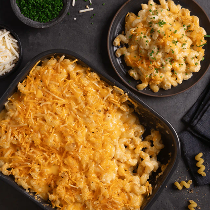 A black baking dish filled with saucy macaroni and cheese and shredded cheese on top. Next to it is a smaller black plate with a serving scooped onto it, topped with chives.