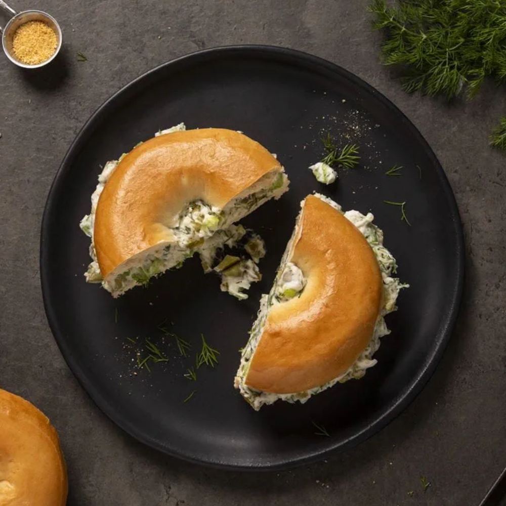 Two halves of a bagel filled with a green herb and Violife Just Like Cream Cheese spread, served on a large black plate on a dark gray surface with dill and a small bowl of seasoning nearby.