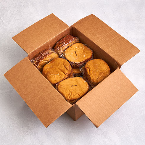 An open brown cardboard box containing several individually wrapped packs of Tofurky Hickory Smoked Deli Slices, photographed from above on a light gray textured surface.