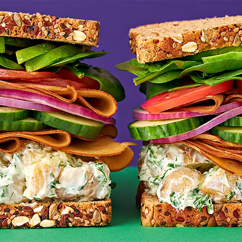 Two tall stacked sandwiches on seeded bread, filled with potato salad, cucumber, red onion, tomato, avocado, and Tofurky Hickory Smoked deli slices, set against a purple background with a green surface.