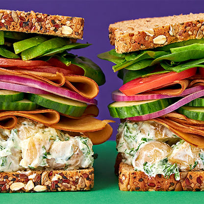 Two tall stacked sandwiches on seeded bread, filled with potato salad, cucumber, red onion, tomato, avocado, and Tofurky Hickory Smoked deli slices, set against a purple background with a green surface.