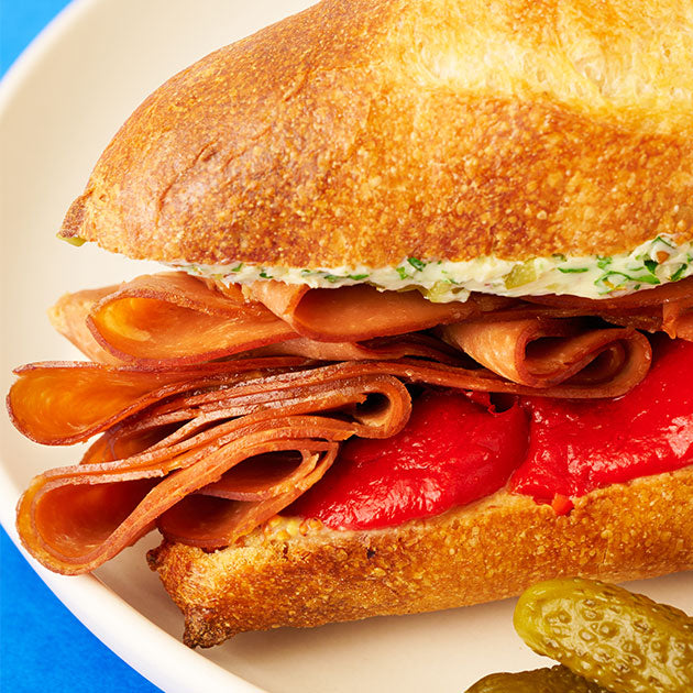 A sliced baguette  sandwich with Tofurky Smoked Ham deli slices and a red sauce, next to some small pickles